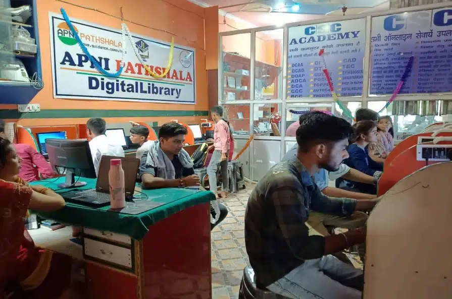 A bustling scene inside a digital library at "Apni Pathshala." Several students are seated at computer stations, working intently. The room is adorned with colourful decorations, and a prominent banner reads "Apni Pathshala Digital Library." To the right, signage for "CSC Academy" lists various courses available. The atmosphere is lively and focused, indicating an active learning environment with people of different ages engaged in digital education.”