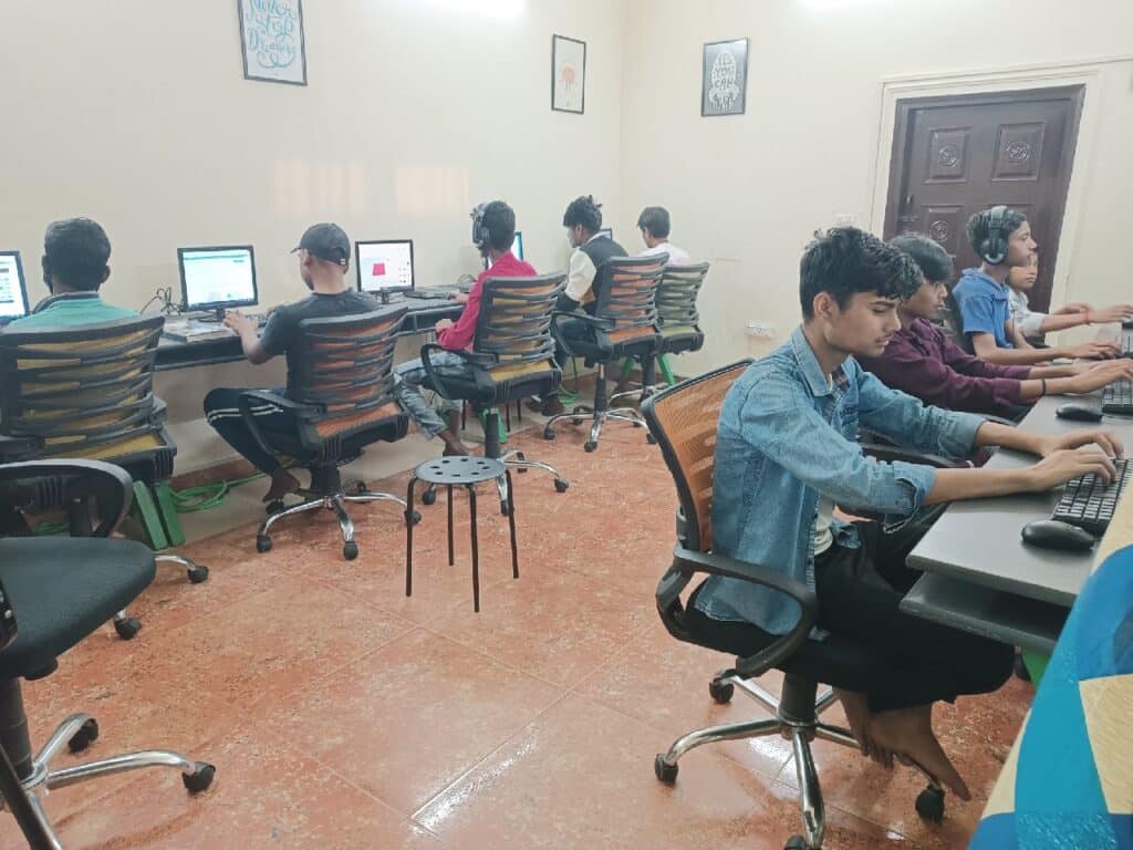 An illustration of an image shows a group of children sitting at desks with computers in a room, seated at computer stations, working intently. The room is adorned with colourful decorations