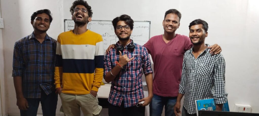 A group of five young men standing together, smiling and posing for the camera in a casual indoor setting with Abhinav standing in the centre and smiling. There is a whiteboard in the background with text written on it.